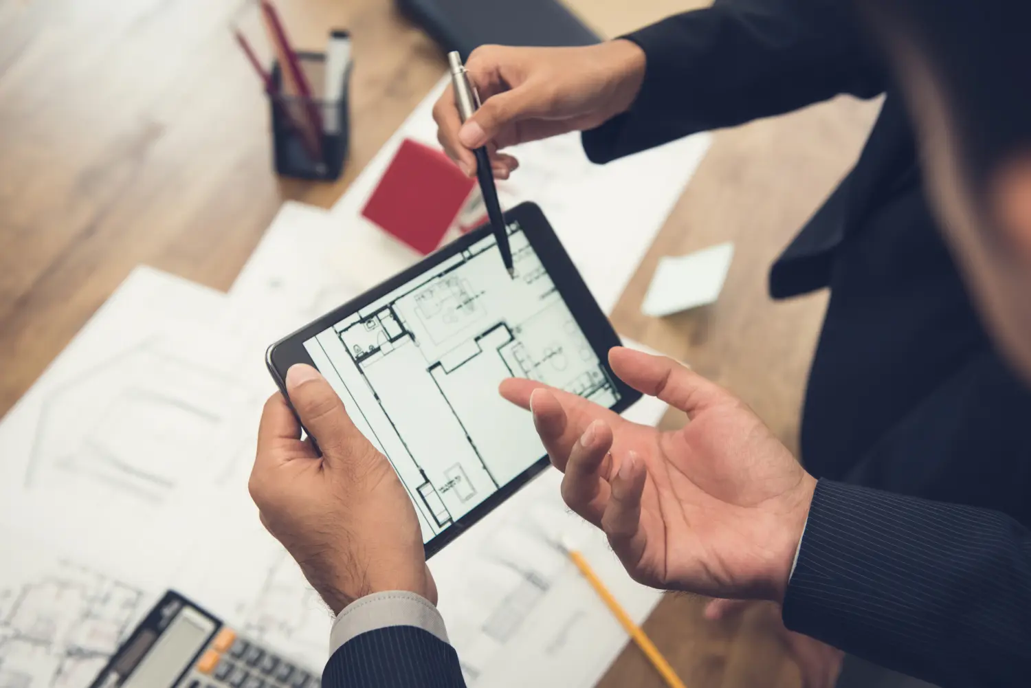 Immobilienmakler mit Kunden- oder Architektenteam, das ein Wohnungsmodell und seine Baupläne digital mithilfe eines Tablets überprüft